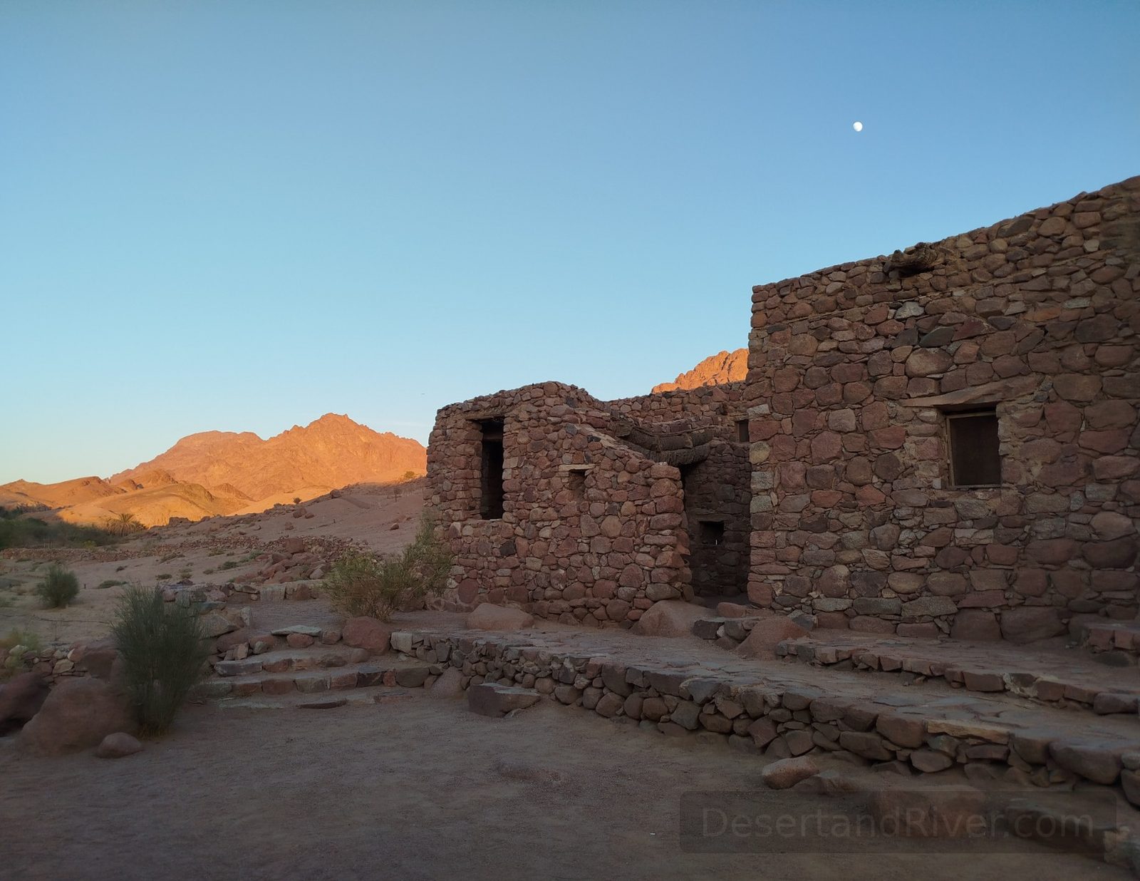 Stone-built Al Karm Ecolodge in South Sinai at sunset, with open terraces and desert mountains under a clear blue sky.