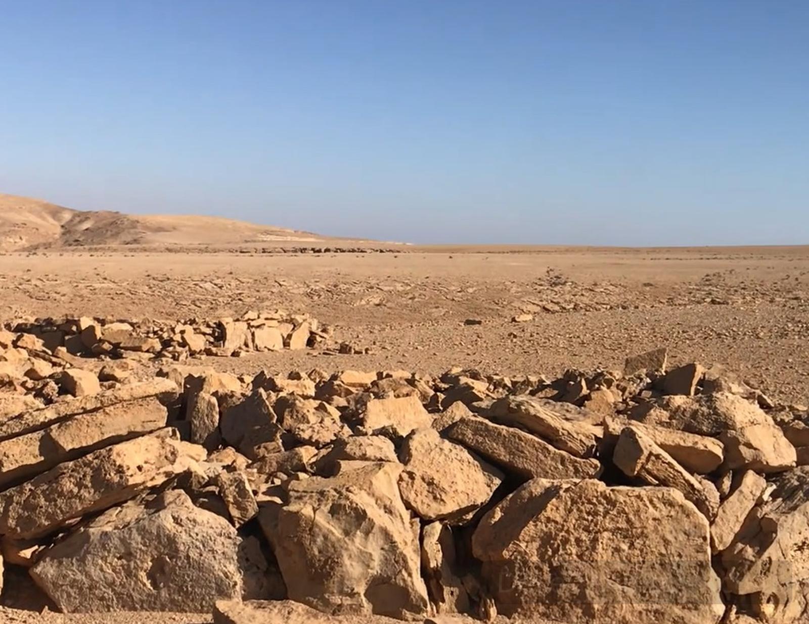 Rock-strewn desert plain at Gebl Gunna in central Sinai, with low stone walls and a wide sandstone mountain ridge under a clear blue sky.