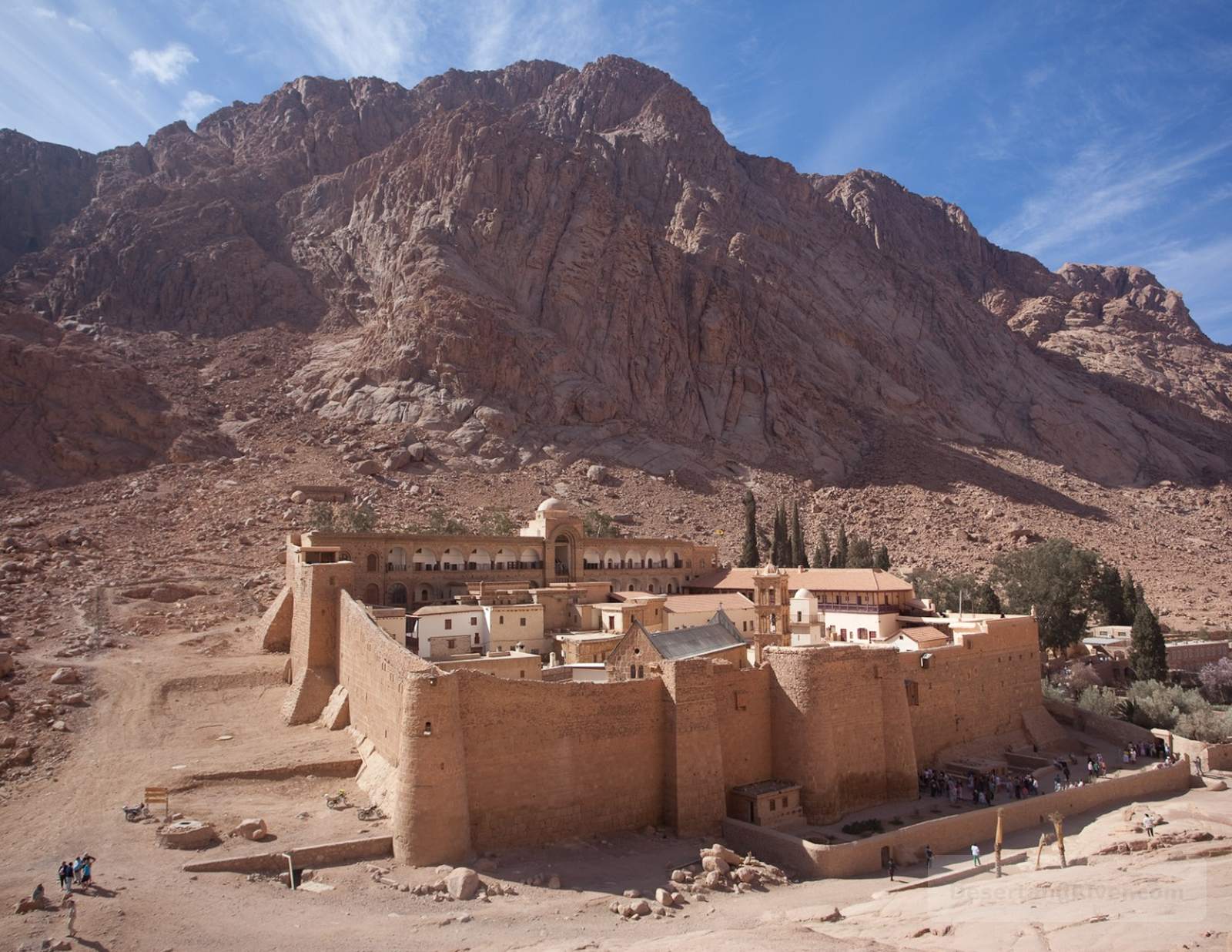 St. Katherine Monastery at the foot of Mount Sinai, a fortified stone complex set against rugged desert mountains under a blue sky.
