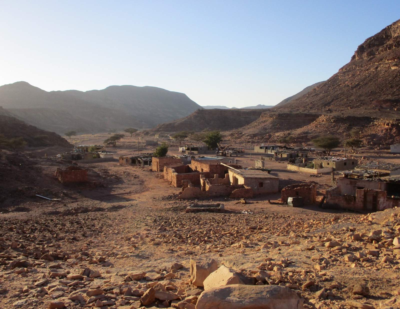Bedouin village in Wadi el Sahu, South Sinai, with stone buildings scattered across a wide desert valley between rocky hills at sunset.
